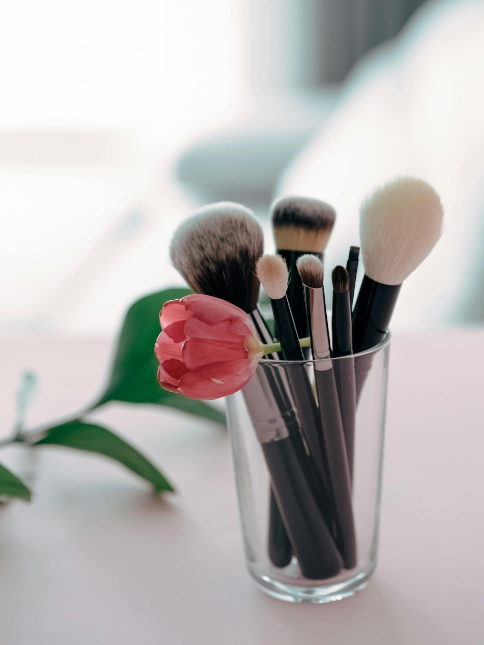 Glass cup holding black-handled makeup brushes with soft bristles and a pink tulip on a light surface, representing elegance and simplicity — a perfect visual for Top Amazon Beauty Finds for Canadian Women